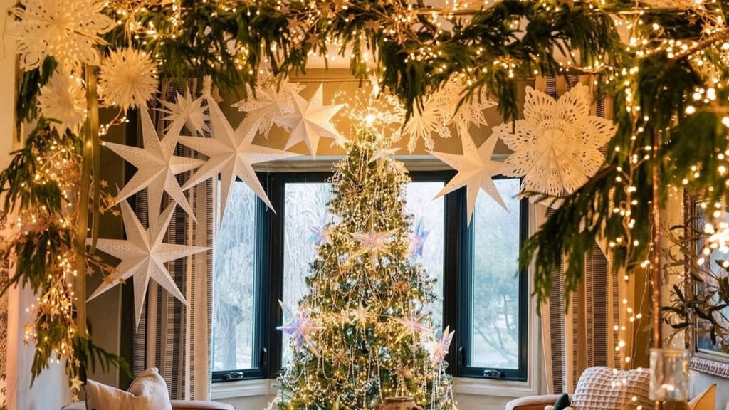 White paper stars and snowflakes hang from an evergreen garland wrapped in twinkle lights over a wide doorway, framing a glowing Christmas tree in front of a black-framed bay window in a warm living room.