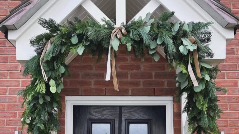 Full pine and eucalyptus Christmas garland draped over a white porch gable on a red brick house, with cream and tan ribbons trailing down and both ends dropping toward a black front door.