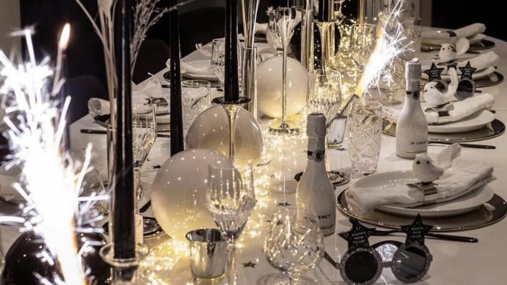 Long white New Year’s Eve table with lit sparklers in the foreground, tall black taper candles, glowing white orbs and fairy lights down the center, mini champagne bottles and star-shaped party glasses at each place setting.