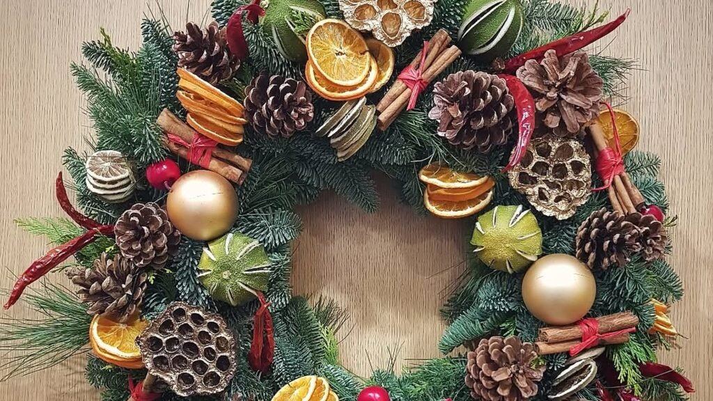 Round Christmas wreath on a light wood door made of fir branches, dried orange slices, cinnamon sticks, pinecones, lotus pods, dried limes and two gold ornament balls, with red chili peppers tucked between.