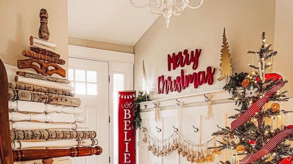 Farmhouse Christmas entryway with a tree stacked from salvaged wooden spindles on the left, a red Merry Christmas sign above a white paneled hook wall, and a lit evergreen tree wrapped in red striped ribbon with dried orange slices on the right.