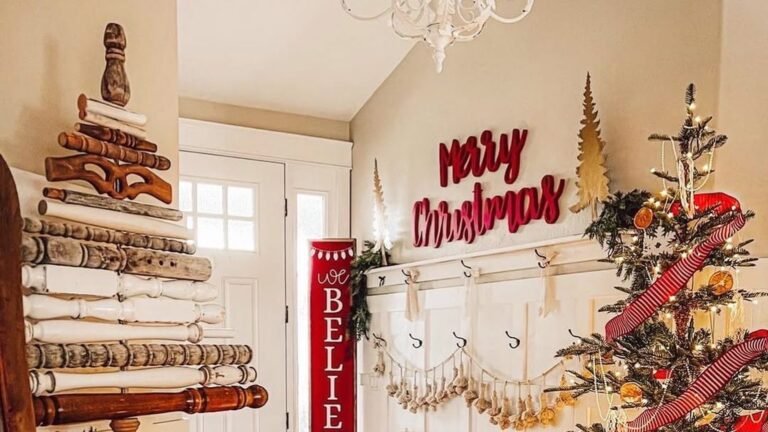 Farmhouse Christmas entryway with a tree stacked from salvaged wooden spindles on the left, a red Merry Christmas sign above a white paneled hook wall, and a lit evergreen tree wrapped in red striped ribbon with dried orange slices on the right.