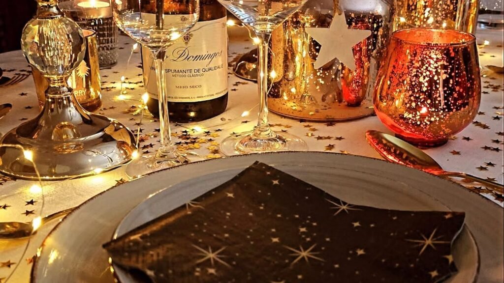 Black napkin printed with tiny gold stars resting on a gray plate, surrounded by scattered gold star confetti, warm fairy lights, a copper mercury glass votive, and a sparkling wine bottle on a white New Year’s Eve table.