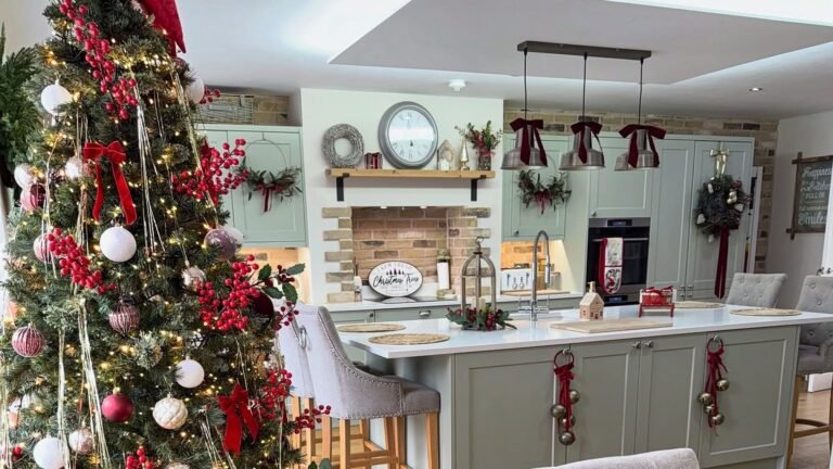 Sage green Christmas kitchen with red velvet bows tied on three pendant lights, jingle bells hanging from red ribbons on the island doors, wreaths on the cabinets, and a tall decorated tree with red berries and warm lights beside the island.