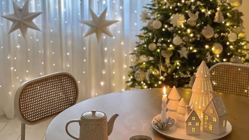Round wooden table with a stoneware teapot and a tray of glowing white ceramic houses and a lit candle, in front of a decorated Christmas tree and white paper stars hanging before a curtain of fairy lights.