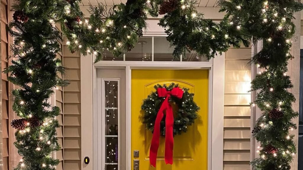 Bright yellow front door with a green wreath and a long red velvet bow, framed by lit evergreen garland with pinecones and warm white lights running down both sides and across the top of the doorway.