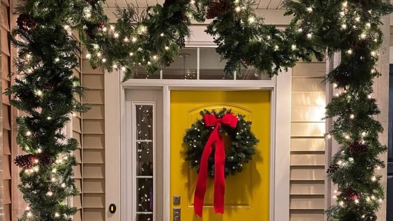Bright yellow front door with a green wreath and a long red velvet bow, framed by lit evergreen garland with pinecones and warm white lights running down both sides and across the top of the doorway.